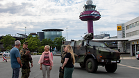 Personengruppe im Gespräch vor einem militärischen Fahrzeug, Tower im Hintergrund