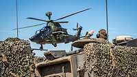 Ein Kampfhubschrauber Tiger fliegt über getarnte Panzerfahrzeuge.