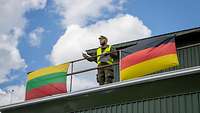 Soldat in gelber Warnweste steht auf Gebäude. Neben ihm eine deutsche und eine litauische Flagge.