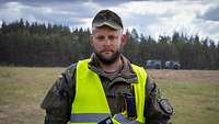 Porträt eines Soldaten in gelber Warnweste in offenem Gelände. Er hält ein Klemmbrett.