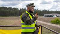 Ein Soldat in gelber Warnweste steht an einem Geländer. Er hält ein Funkgerät und ein Klemmbrett.