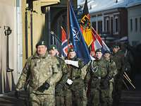 Mehrere Soldaten unterschiedlicher Nationen marschieren mit Flaggen zwischen Häusern.