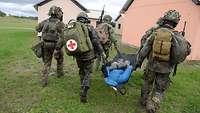 Mehrere Soldaten tragen ein Rettungstuch mit Übungspuppe. Einer trägt ein Rucksack mit rotem Kreuz.