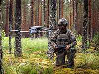 Eine Drohne schwebt im Wald, im Hintergrund ist der zugehörige Soldat mit Fernsteuerung zu sehen.