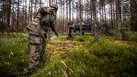 Soldaten bauen eine Antenne im Wald auf, im Hintergrund steht ein abgetarntes Transportfahrzeug.