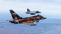 Ein grauer Kampfjet fliegt hinter einem Jet mit deutschen Hoheitszeichen.