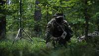 Ein Soldat kniet im Wald und arbeitet an einem Gerät. Neben ihm steht eine Antenne.