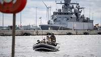 Soldaten fahren in einem Schlauchboot im Hafen. Im Hintergrund liegt ein Kriegsschiff an der Pier.