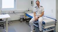 Ein Mann in Arztuniform sitzt auf einer Liege und lächelt. 