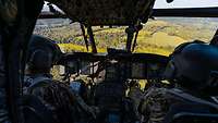 Zwei Piloten sitzen im Cockpit eines Hubschraubers. Draußen sieht man Felder und Wald.
