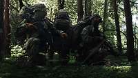 Drei Soldaten knien gut getarnt in einem Waldstück.