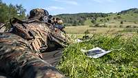A soldier lies on a meadow with a machine gun at the ready.
