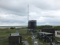 Eine große Radarantenne auf einem Fahrzeug, im Vordergrund Container, im Hintergrund das Meer