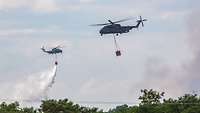 Zwei Militärhubschrauber fliegen mit Behältern zum Löschen von Bränden über Baumen