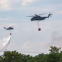 Zwei Militärhubschrauber fliegen mit Behältern zum Löschen von Bränden über Baumen