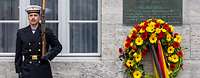 Ein Soldat steht an einer Wand neben einer Gedenkplatte unter der ein Kranz mit Blumen hängt