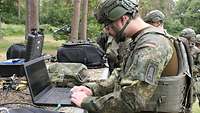 Ein Soldat mit Helm und Kampfweste arbeitet im Wald an einem Laptop.