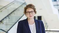 Porträt einer Frau mit kurzem Haar und Brille. Sie trägt einen blauen Blazer und eine silberne Kette