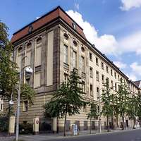 Ein mehrstöckiges historisches Gebäude mit Sandsteinfassade, umgeben von Bäumen und Laternen.