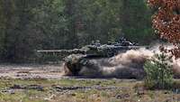 Ein getarnter Kampfpanzer Leopard fährt schnell durchs Gelände, Sand wirbelt auf.
