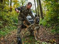 Ein Soldat steht mit einem angeleinten Hund auf einem Waldweg, im Hintergrund ein Fahrzeug