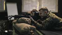 Ein Soldat mit zugekniffenem Auge liegt auf einem Tisch, die Waffe ist auf Sandsäcken aufgelegt.