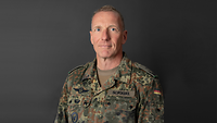 Ein Soldat im Feldanzug vor einer dunklen Leinwand schaut in die Kamera.