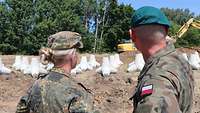 Eine deutsche Soldatin und ein polnischer Soldat sind von hinten zu sehen, vor ihnen liegen Betonsperren