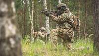 Ein Soldat kniet mit einer Waffe im Anschlag in einem Waldgebiet.