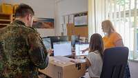 Ein Soldat steht im Büro und schaut auf ein Tablet, während zwei Zivilistinnen an einem PC arbeiten.