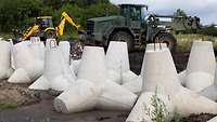 Mehrere weiße Betonpanzersperren stehen im Schlamm vor einem grünen Radlader