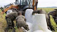 Drei Soldaten in verschiedenen Flecktarnuniformen platzieren weiße Betonpanzersperren