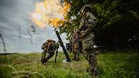 Soldaten stehen im Gelände an einem Mörser und schießen. Ein Feuerball ist zu sehen.