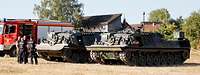 Zwei Bergepanzer der Bundeswehr stehen neben einem Löschfahrzeug auf einer Freifläche vor Häusern.