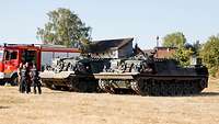 Zwei Bergepanzer der Bundeswehr stehen neben einem Löschfahrzeug auf einer Freifläche vor Häusern.