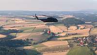 Ein Hubschrauber mit Löschwasserbehälter in der Luft über einer Landschaft.