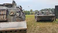 Zwei Panzer stehen auf einer Freifläche. Im Hintergrund fliegt ein Hubschrauber mit Löschbehälter.