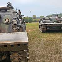 Zwei Panzer stehen auf einer Freifläche. Im Hintergrund fliegt ein Hubschrauber mit Löschbehälter.