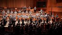 Ein Orchester steht in einem Musiksaal auf einer Bühne. Vor der Bühne stehendes Publikum