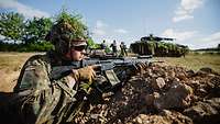 Soldaten mit Waffen stehen in einer Stellung. Im Hintergrund sind Soldaten und ein Panzer zu sehen.
