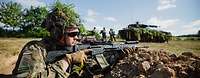 Soldaten mit Waffen stehen in einer Stellung. Im Hintergrund sind Soldaten und ein Panzer zu sehen.