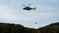 Ein Hubschrauber vom Typ NH-90 mit einem Löschwasserbehälter fliegt über ein Waldgebiet.