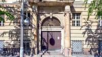 Ein mehrstöckiges historisches Haus mit einem großen verzierten Eingangstor. Rechts und links davon stehen zwei Bäume.