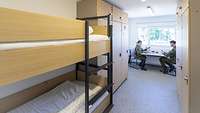 Blick in eine Unterkunft mit Doppelstockbett, Spinden und einem Tisch, an dem zwei Personen sitzen.