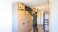 Blick in eine Unterkunft. Ein Soldat holt Kleidung aus dem Aufbauschrank über dem Spind.