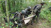 Vier Soldaten im Wald tragen eine verletzte Person in einer Trage.