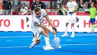 Ein Deutscher Hockeyspieler schlägt einen Ball. Dabei wirbelt er Sand vom blauen Kunstrasen auf. 