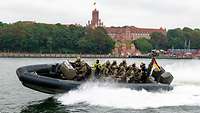 Ein Schlauchboot mit Polizisten fährt über ein Gewässer. Am Ufer steht ein historisches Gebäude.