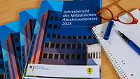 Auf einem Tisch liegen mehrere Jahresberichte. Daneben liegen eine Brille, ein Füller und ein Schreibblock.