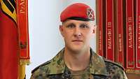 Ein Soldat mit eine roten Barett steht vor einer Truppenfahne.